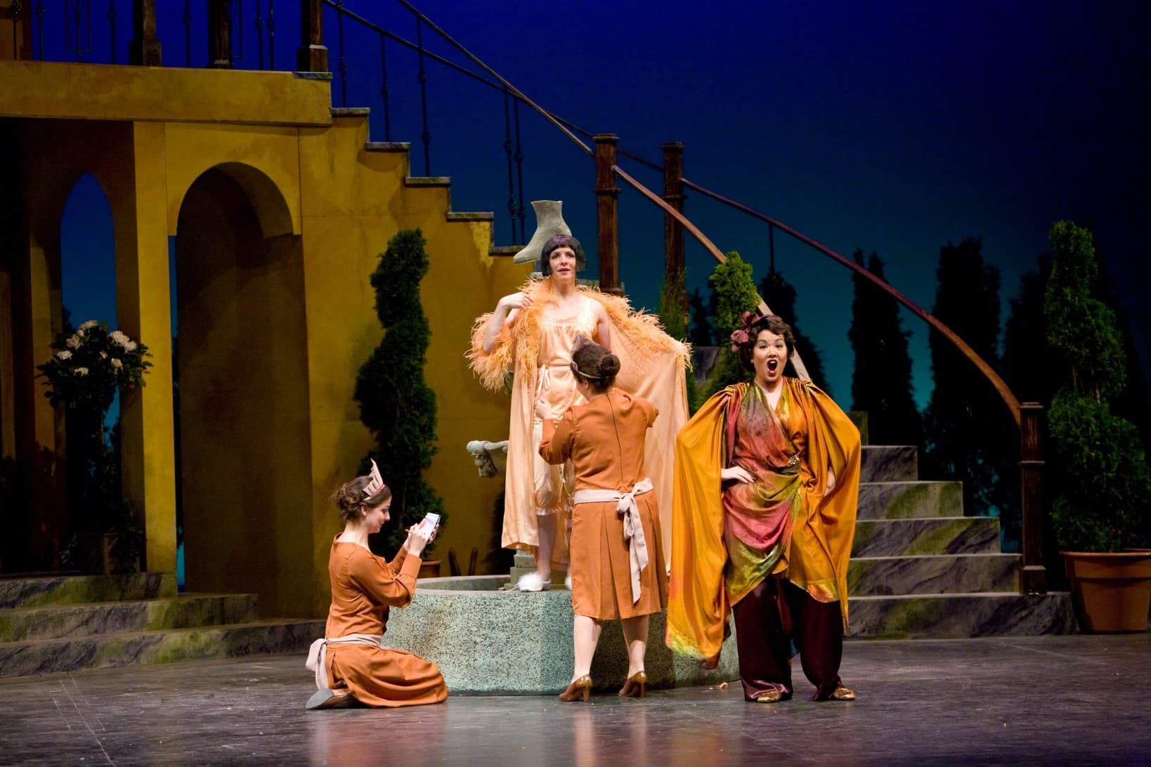 This image contains the same characters as the other photos. While the characters in the other photos were consistently dressed
in each picture, they are wearing more decorative clothes in this scene. Sarah is standing on the fountain with a feathered
boa, while the woman who wear red is standing in front of her in a yellow cape and singing.