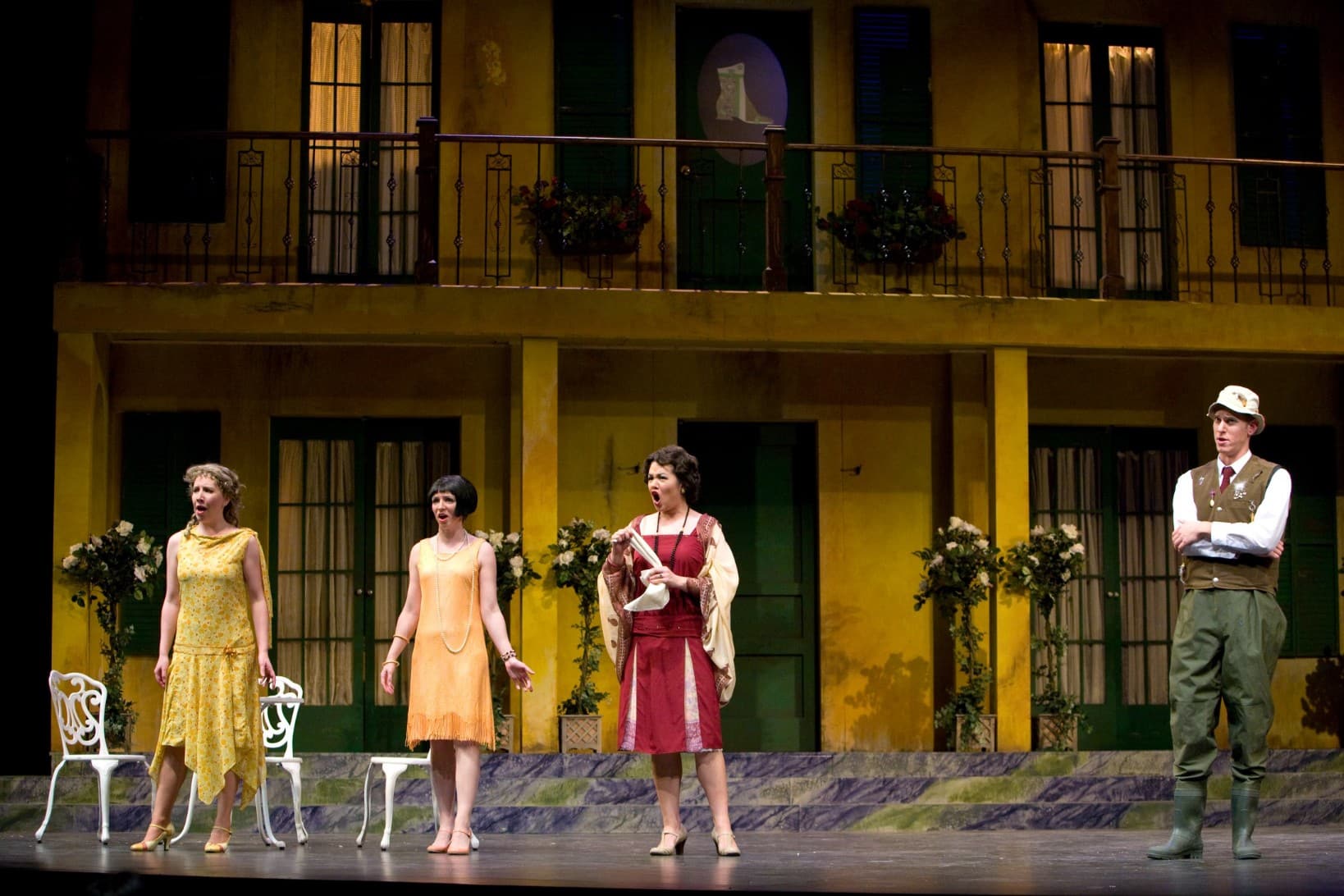 Three women singing in unison on stage, two in yellow dresses and one wearing red. A man on stage in a vest watches on the
side.
