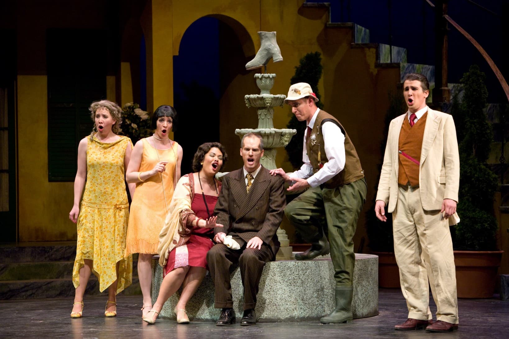 6 people on stage, singing in unison. Two are seated on a fountain while the rest are standing around them.