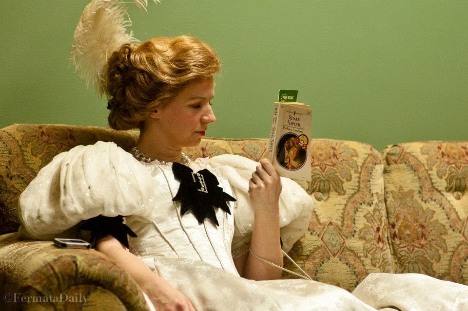 A woman is wearing a turn of the century dress with large poofy shoulder pads, a corset, and a plume in her hair. She is
reading the back of a book.