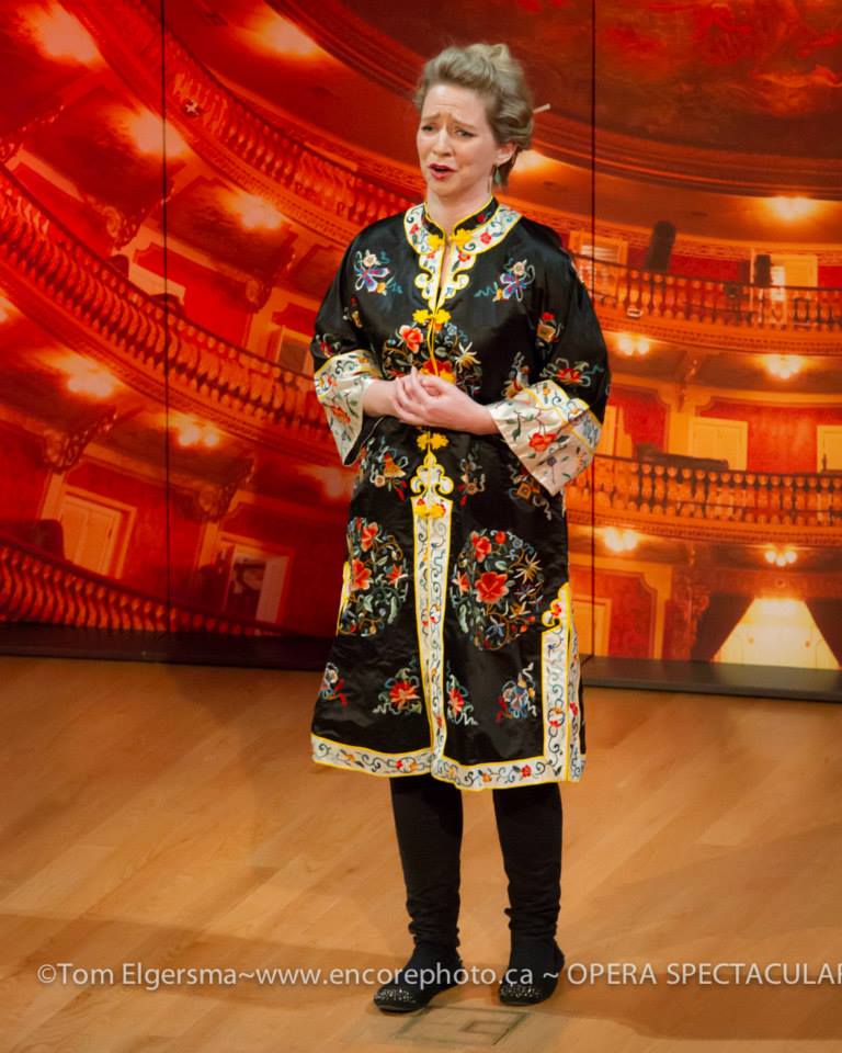 Sarah is standing on stage, wearing a frock in a traditional Chinese style with gold sleeves and lots of embroidery.