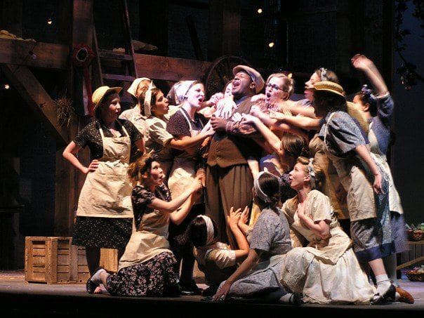 A man is center stage, singing to the sky, and around him there are around 10 women singing with him, propping him up, and
embracing him in an uplifting manner. The light is shining directly in his face, giving the scene a look of hope for the
future.