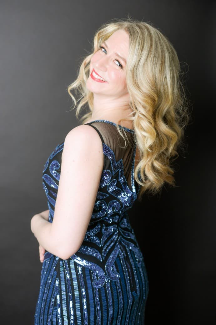 Sarah is wearing a blue sequins dress that has black mesh around the shoulders. Her hair is in curls and she is looking over
her shoulder at the camera.