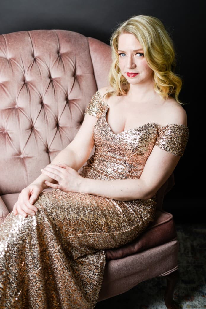 Sarah is in her gold sequins dress and sitting on the pink velvet couch. The dress has sleeves that go off the shoulder,
and the length goes down to her feet.