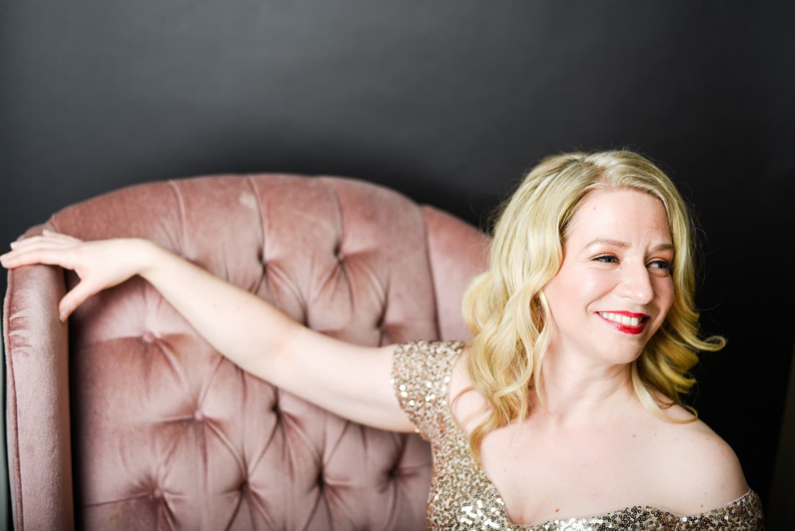 Sarah is in the gold sequins dress and sitting on the couch. she has her right arm on the couch, while her body is centered
and her face is looking to her left.