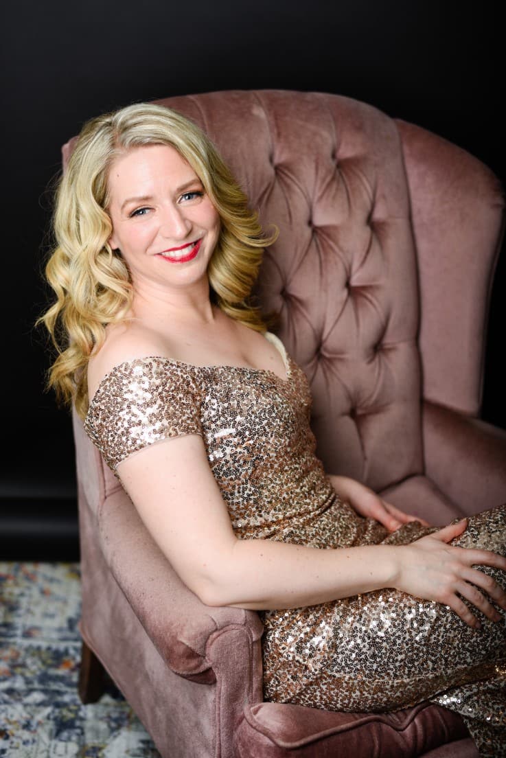 Sarah is sitting on the pink velvet couch in her gold sequins dress. There is a faded colourful carpet on the floor, and
she is looking at the camera with her body facing away to the right of the frame.