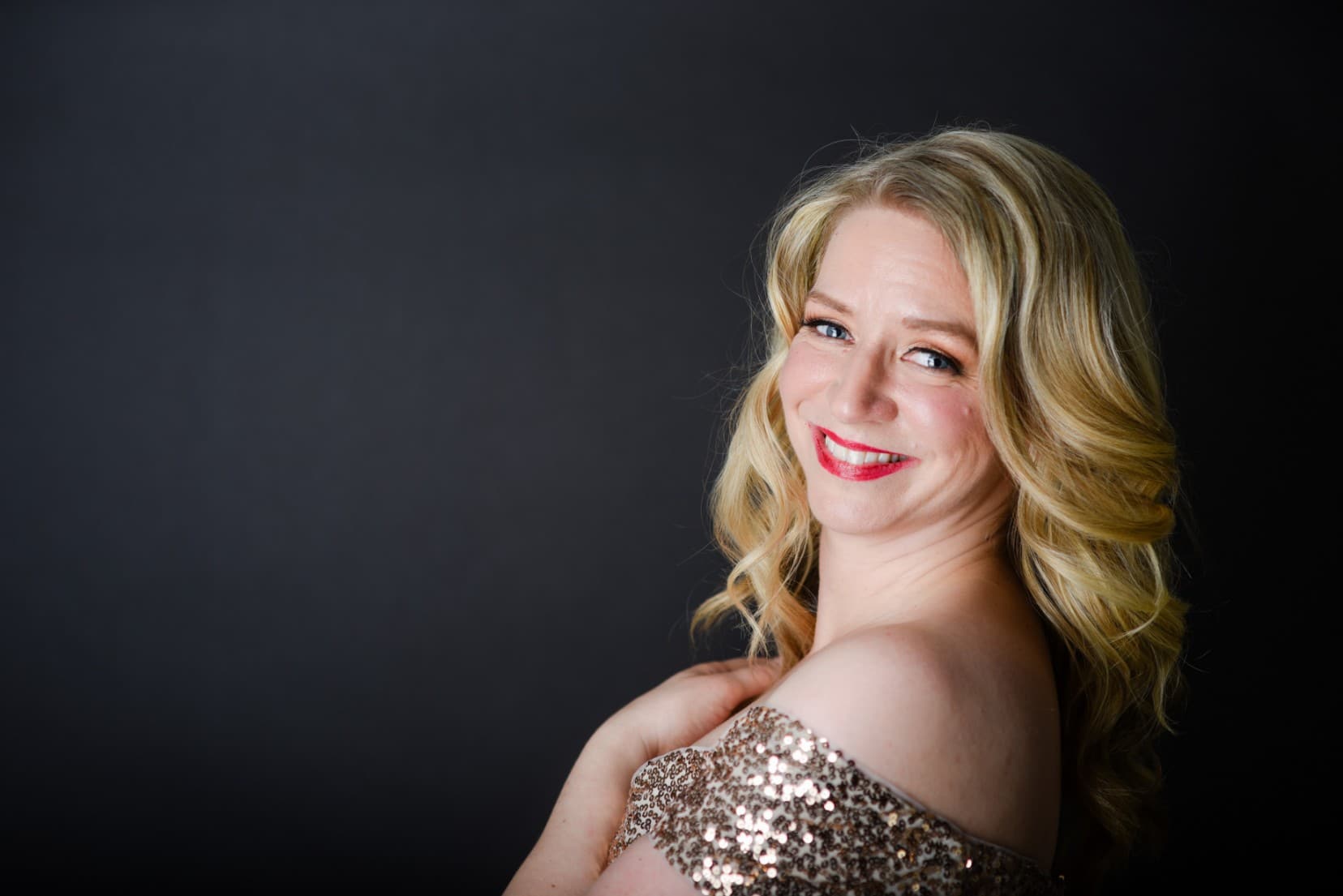 Sarah is wearing her sequins dress and looking over her shoulder with on hand on her heart.