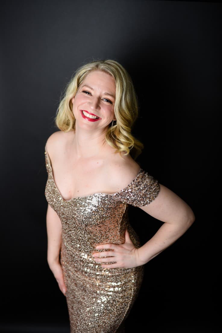 Sarah is wearing her gold sequins dress. The camera is higher than her. Her left hand is on her waist and she is smiling
up at the camera.