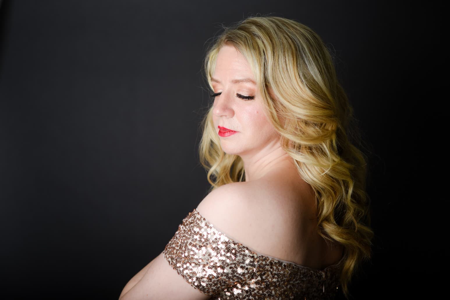 Sarah is wearing a gold sequins dress. The image just shows above her shoulders, and she is turned away from the camera.
Her face is turned slightly back, and she is looking down with soft eyes.